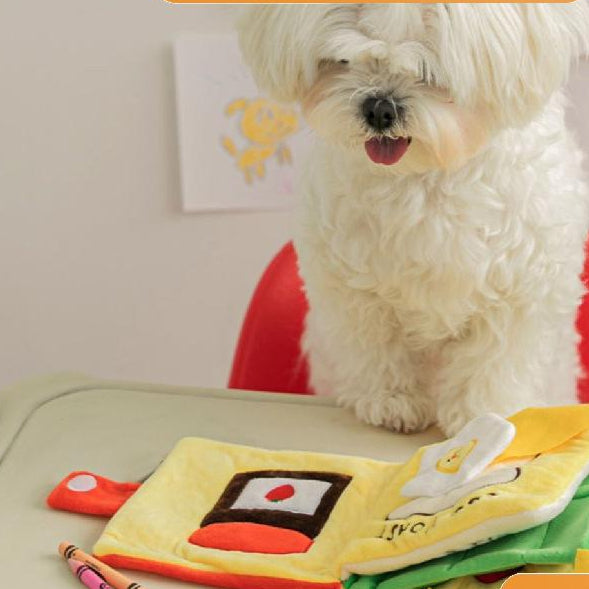 White dog sitting at a table with colorful children's books and crayons on a light-colored surface.