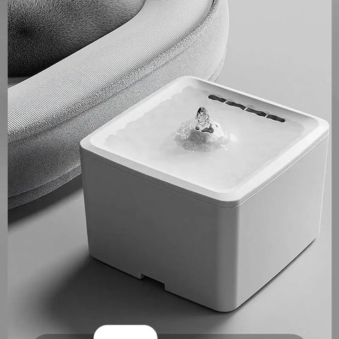 White square automatic pet water fountain placed next to a plush grey pet bed.