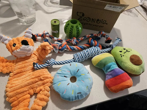 Assortment of plush dog toys including a blue donut, green avocado, orange fox, rope toys, and green waste bags on a counter, customer review image.