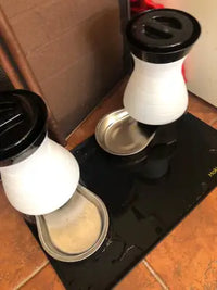 Customer review image of two white and black automatic pet feeders and water fountains with stainless steel bowls set on a waterproof black silicone feeding mat with a raised lip.