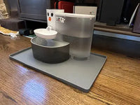 Automatic plastic pet water fountain with a built-in reservoir sitting on a raised-edge gray silicone spill-proof mat on a hardwood floor.