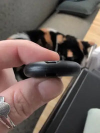 A side profile of a black smart Bluetooth tracker held in fingers with a Calico cat resting in the blurred background.