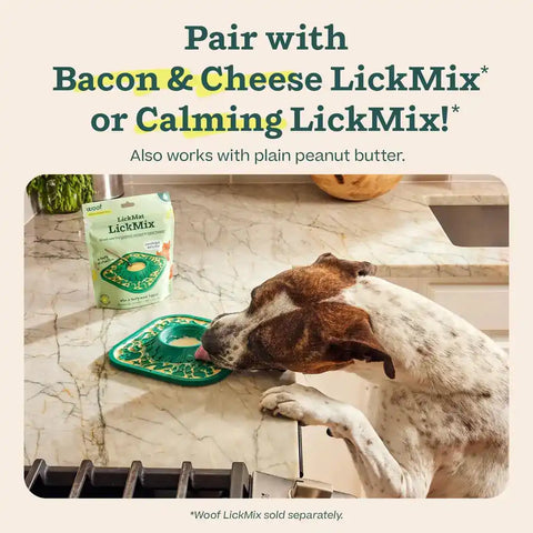 Dog licking treat spread from green LickMat next to a bag of Woof LickMix on a kitchen counter.