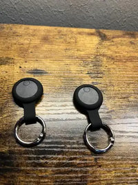 Two round black smart pet trackers inserted into silicone keychain cases placed on a rustic wooden tabletop.