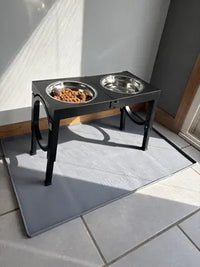 Black elevated dog bowl stand with two stainless steel bowls filled with dry kibble, placed on a gray silicone floor protector mat.