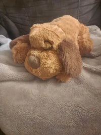 A happy Golden Retriever dog laying on a colorful patterned bed cover with its paws resting over a brown plush puppy toy.
