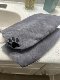 Two folded grey microfiber dog drying towels featuring a black embroidered paw print detail, sitting on a white speckled bathroom counter next to a sink.