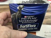A person holding an open box of Purina Pro Plan FortiFlora canine probiotic supplements revealing the silver sachets inside.