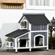 Grey tabby cat looking out from the upper window of a multi-level grey and white outdoor wooden cat house.