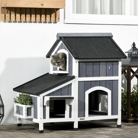 Grey tabby cat looking out from the upper window of a multi-level grey and white outdoor wooden cat house.