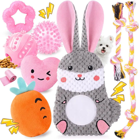 A cute assortment of pink puppy toys featuring a large grey bunny plush, a pink smiling heart, a carrot toy, pink teething rings, a treat ball, rope toys, and pink poop bags.