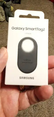 A person holding the retail packaging for a black Samsung Galaxy SmartTag2 1-pack.