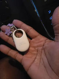 A white Samsung SmartTag2 attached to a decorative silver keychain with purple flower charms held in a hand.