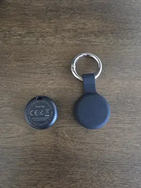 The back of a SeekTag smart finder showing model information next to a black silicone keychain holder on a wooden surface.