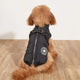 Small dog wearing a black puffer vest with a logo on a wooden floor.