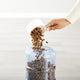 Person pouring dry dog food into the top-loading reservoir of a white gravity pet feeder.