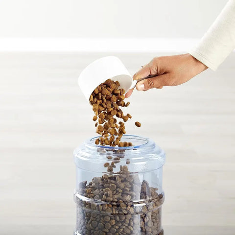 Person pouring dry dog food into the top-loading reservoir of a white gravity pet feeder.