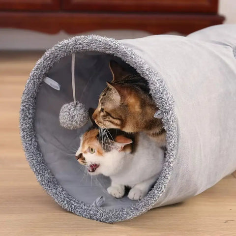 Two cats playing with a plush tunnel toy on wooden floor.