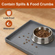 Grey silicone pet food mat with raised edges containing water spills and dry kibble crumbs next to a stainless steel dog bowl.