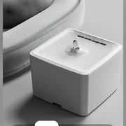 White square automatic pet water fountain placed next to a plush grey pet bed.