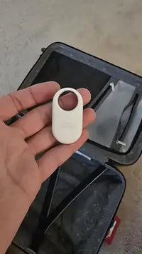 A person holding a white Samsung SmartTag2 tracker over an open suitcase to demonstrate its size.